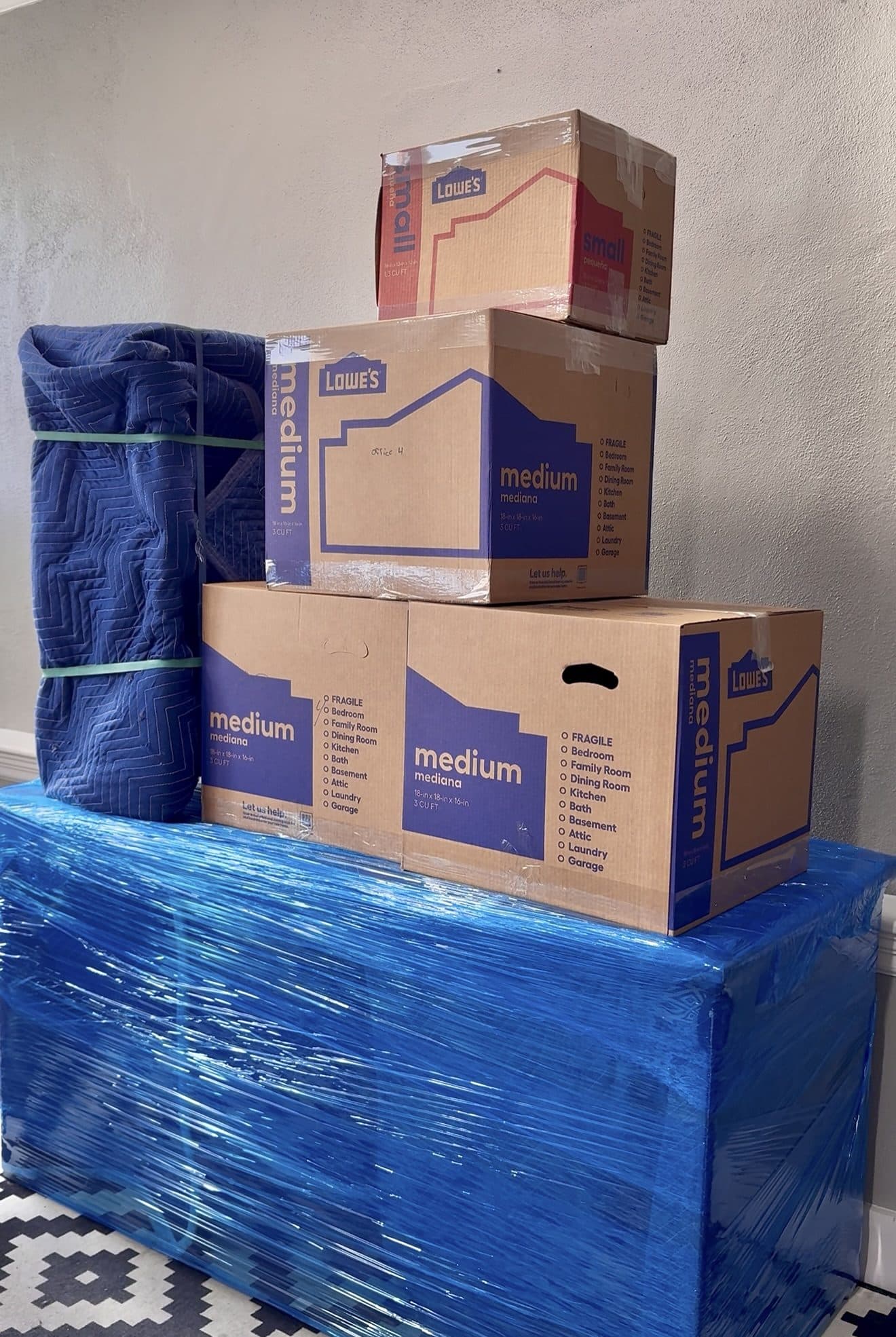 Labelled moving boxes stacked beside a moving blanket and a shrink-wrapped dresser