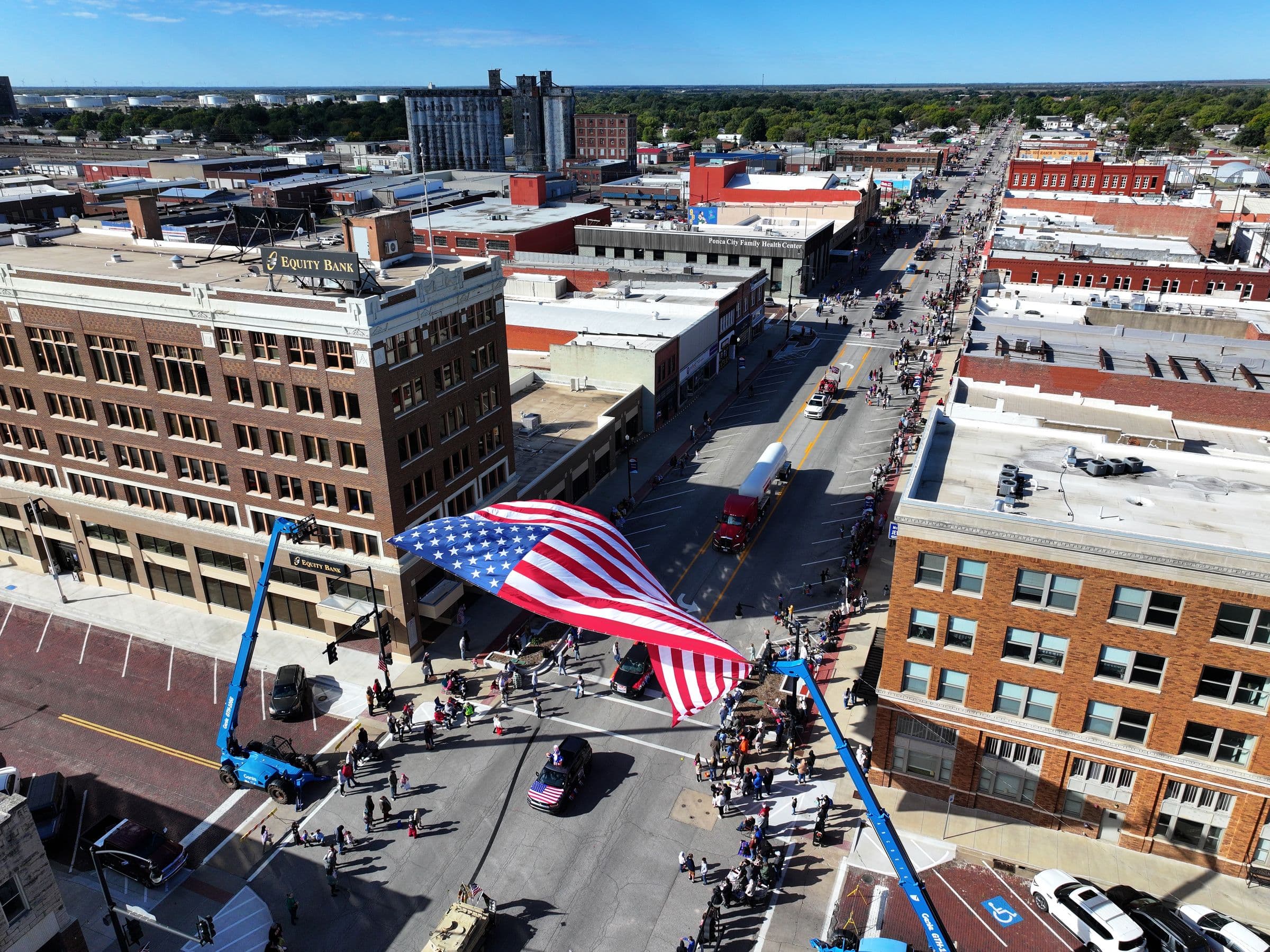 A parade on Grand Avenue in downtown Ponca City, with a large American flag suspended over the street